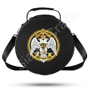Könnte beinhalten: Schwarze runde Tasche mit Griff und Schultergurt. Die Tasche hat ein gold-weißes Emblem mit einem doppelköpfigen Adler, einer Krone und der Zahl 33. Das Emblem ist von einem goldenen Rand umgeben.
