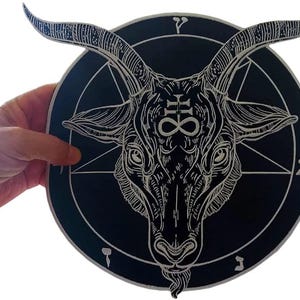 Op de afbeelding: Een zwart, cirkelvormig decoratief stuk met een gedetailleerde witte illustratie van een geitenkop met hoorns, een pentagram en andere symbolen. Het kunstwerk wordt door een hand vastgehouden, waarbij het gedetailleerde ontwerp en de contrasterende kleuren worden benadrukt.