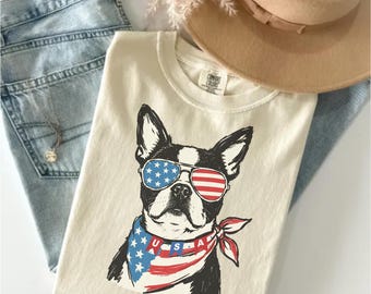 May include: A cream-colored t-shirt featuring a graphic of a Boston Terrier wearing sunglasses with a stars and stripes pattern and a matching American flag bandana with the text USA. The shirt is paired with light blue denim jeans and a tan hat.