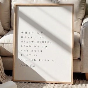Puede incluir: Una impresión enmarcada con un marco de madera clara. El fondo blanco presenta texto negro que dice: "Cuando mi corazón está abrumado: llévame a la roca que es más alta que yo". El texto "Salmo 61:2" está debajo.