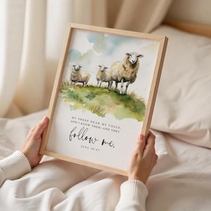 May include: Framed watercolour print depicting three sheep on a green hill. The text reads: "My sheep hear my voice, and I know them, and they follow me. John 10:27." The frame is made of light-colored wood.