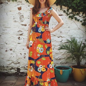 May include: A woman in a long, sleeveless orange dress with a vibrant floral design, a straw hat, and sunglasses. The dress has a ruffled neckline and hem. The background shows a white wall with climbing plants and potted plants.