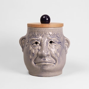 Könnte beinhalten: Ein Keramiktopf mit Gesichtsdesign, mit einem Holzdeckel und einem dunkelvioletten Knopf. Der Topf ist hellviolett mit einem weinenden Gesichtsdesign, einschließlich Tränen. Der Topf hat zwei ohrähnliche Griffe an den Seiten.