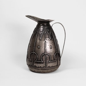 Può includere: Una brocca in metallo argentato con manico curvo e beccuccio. La brocca ha un corpo arrotondato con motivi decorativi e rivetti. Lo sfondo è bianco.