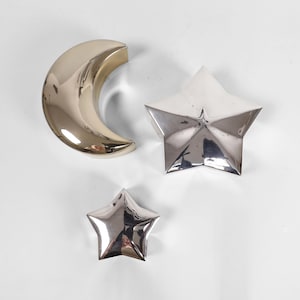 Könnte beinhalten: Drei metallische Himmelskörper: ein Halbmond, ein fünfzackiger Stern und ein kleinerer Stern. Der Mond hat eine goldene Oberfläche, während die Sterne silberfarben sind. Alle drei Objekte haben eine reflektierende, polierte Oberfläche.