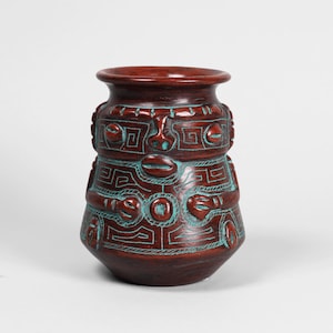 Puede incluir: Un jarrón de cerámica marrón con detalles turquesa. El jarrón presenta un diseño detallado de formas geométricas y caras estilizadas. El recipiente tiene un cuerpo redondeado y un borde ligeramente ensanchado, mostrando una estética artesanal única.