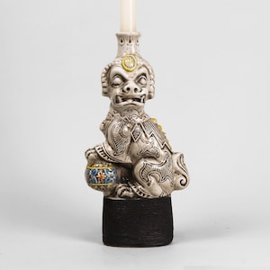 May include: A ceramic candle holder featuring a detailed, seated mythical creature with a fierce expression. The holder is primarily off-white with gold and blue accents, and a black base. A white candle is inserted in the top.
