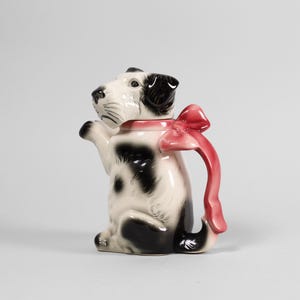 Könnte beinhalten: Eine Keramik-Teekanne in Form eines sitzenden Hundes mit schwarzen Flecken und einer roten Schleife. Der Hund hat schwarze Ohren und ein weißes Gesicht. Griff und Ausguss sind in das Design integriert, was ein skurriles Stück ergibt.