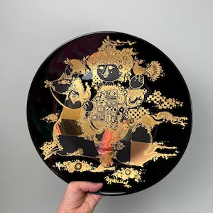 Puede incluir: Un plato de cerámica negro con un diseño dorado de una persona montando a caballo. El diseño es intrincado y detallado, con muchos patrones y formas pequeñas. El plato está firmado "Tapio Wirkkala" en oro.