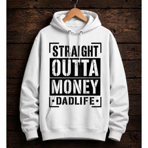 May include: White hoodie featuring the text "STRAIGHT OUTTA MONEY DADLIFE" in black. The sweatshirt has a drawstring and a front pocket. It is displayed on a wooden hanger against a dark wood backdrop.