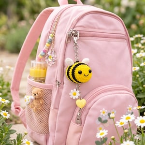 Può includere: Uno zaino rosa con un portachiavi a forma di ape all'uncinetto, un ciondolo di perline e un piccolo orsacchiotto nella tasca laterale. Il portachiavi a forma di ape è giallo e nero con ali bianche. Una bottiglia di succo è nella tasca laterale.
