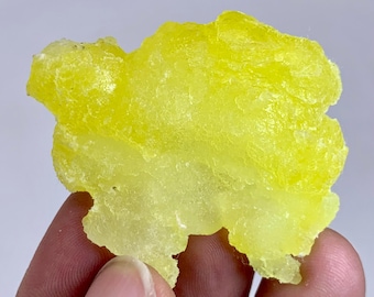 May include: A bright yellow, irregularly shaped mineral specimen. The mineral has a textured surface and a translucent appearance, with variations in color intensity. The specimen is held between fingers, showcasing its natural form and color.