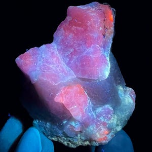 May include: A large, pink and white crystal with a rough, natural texture. The crystal glows under UV light, showing a vibrant pink hue.
