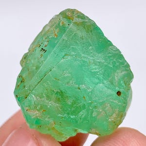 May include: A raw, uncut green fluorite crystal. The gemstone has a crystalline structure with a vibrant, translucent green color. The surface has a rough, natural texture with small brown inclusions.