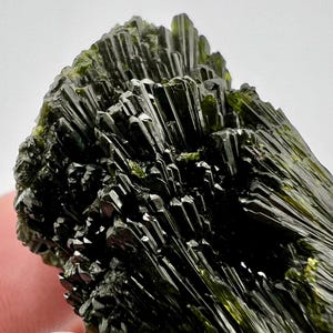 Puede incluir: Un cúmulo de cristales de color verde oscuro, en forma de aguja, con un brillo metálico brillante. Los cristales están dispuestos en un patrón radial, creando una exhibición única y llamativa.