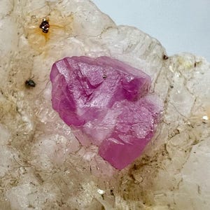 May include: A pink crystal with a rough, faceted surface is embedded in a white, crystalline matrix. The crystal has a translucent appearance and is surrounded by smaller, white crystals.