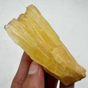 Könnte beinhalten: Ein großer, gelber, durchscheinender Kristall mit einer gestreiften Textur. Der Kristall hat die Form einer langen, dünnen Klinge.