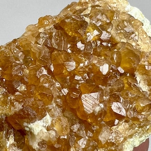 Może przedstawiać: Zbliżenie na skupisko złoto-brązowych kryształów. Kryształy mają fasetowaną, geometryczną strukturę, z ostrymi krawędziami i odblaskowymi powierzchniami. Ogólny wygląd jest szorstki i naturalny, z jasną podstawą.