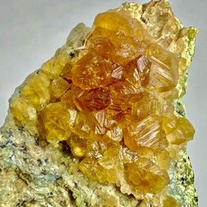 Può includere: Primo piano di un campione minerale con gruppi di cristalli giallo dorato. I cristalli hanno una struttura geometrica e angolare e sono su una matrice di roccia verde e grigia. L'aspetto generale è ruvido e naturale.