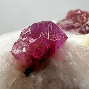 May include: Close-up of a vibrant, faceted, magenta-colored crystal formation. The crystal is set against a backdrop of white, crystalline rock. The image highlights the natural beauty and geological detail of the mineral specimen.