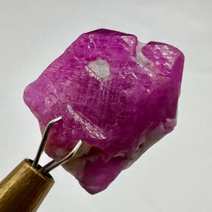 May include: A large, rough, pink ruby crystal with a white inclusion. The crystal is being held by a pair of tweezers.