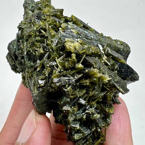Può includere: Primo piano di un grezzo campione di minerale verde scuro. Il minerale ha una struttura cristallina con molti piccoli cristalli aghiformi raggruppati. Il colore varia dal verde scuro a una tonalità verde-giallastra più chiara.
