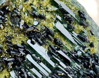 May include: Close-up of a mineral specimen featuring dark green, elongated crystals and smaller, black, and yellow-green crystalline structures. The crystals are clustered together, creating a textured surface. The mineral has a natural, raw appearance.