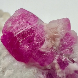 Puede incluir: Un cristal de rubí rosa vibrante con una superficie rugosa y texturizada. El cristal está incrustado en una matriz blanca.