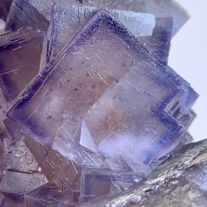 May include: Close-up of a fluorite crystal cluster. The crystal structure features a series of square and rectangular formations with a gradient of colors, transitioning from a pale lavender to a deep purple hue. The crystal has a natural, rough texture.