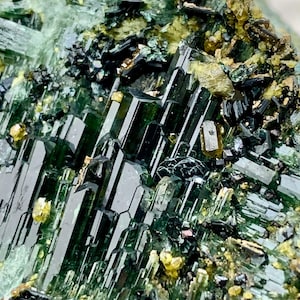 Może przedstawiać: Zbliżenie na okaz minerału z ciemnozielonymi, wydłużonymi kryształami o szklistym połysku. Kryształy są zgrupowane, przeplatane mniejszymi, żółtymi i czarnymi formacjami mineralnymi. Ogólna kompozycja to mieszanka tekstur i kolorów.