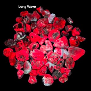 May include: A collection of irregularly shaped, red-colored stones with black and white inclusions, against a black background. The text "Long Wave" is in the upper left corner. The stones have a glowing appearance.