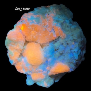 May include: A close-up of a mineral specimen with a black background. The mineral has a textured surface with orange and blue hues. The text "Long wave" is in the upper left corner. The mineral appears to glow.
