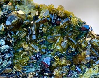 May include: Close-up of a mineral specimen featuring a cluster of lustrous, prismatic crystals. The crystals display a range of colors, including deep greens, yellows, and hints of blue and black, creating a striking visual contrast. The overall texture is rough and crystalline.