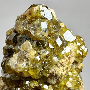 Pode incluir: Close-up de um aglomerado de cristais amarelo-dourados com uma forma geométrica facetada. Os cristais estão agrupados em uma matriz áspera bege. Os cristais têm uma qualidade translúcida, refletindo a luz.
