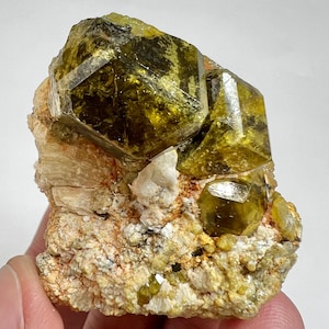 Op de afbeelding: Een cluster van levendige, gefacetteerde, geelgroene kristallen, mogelijk een mineraalmonster, geplaatst tegen een matrix van beige en bruin gesteente. De kristallen hebben een glasachtige uitstraling, met scherpe randen en een kristallijne structuur. De algehele samenstelling is natuurlijk en aards.
