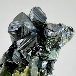 Puede incluir: Primer plano de un espécimen mineral que presenta grupos de cristales geométricos oscuros y metálicos. Los cristales tienen una superficie estriada y reflectante, con toques de color iridiscente. La roca base es una mezcla de tonos verdes y grises, con algunos acentos amarillos.