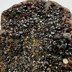 Peut inclure: Gros plan sur un spécimen minéral avec une surface rugueuse et texturée. La surface est recouverte de grappes de petits cristaux sombres et brillants, avec des teintes brun rougeâtre et noires. L'aspect général suggère une pierre précieuse ou un minéral brut naturel.