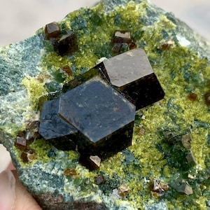 Op de afbeelding: Close-up van een mineraalmonster met donkere, glanzende, geometrische kristallen. De kristallen zijn geplaatst tegen een matrix van groene en geelgroene mineralen, wat een opvallend contrast creëert. De algehele textuur is ruw en natuurlijk.