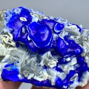 May include: A close-up of a vibrant blue mineral specimen with a glossy, textured surface. The mineral is interspersed with gray and white crystalline formations, creating a striking contrast. The overall appearance suggests a natural, raw gemstone.