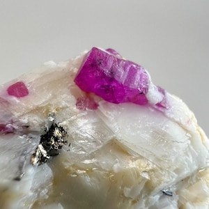 以下が含まれることがあります： 鮮やかなマゼンタ色の結晶が特徴の鉱物標本。 白とクリーム色の岩を背景に、黒と銀の斑点があります。