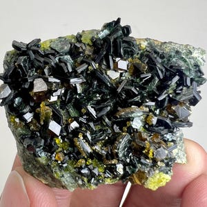 Puede incluir: Primer plano de un espécimen mineral con grupos de cristales oscuros y alargados. Los cristales son de un negro profundo y reflectante, contrastados por áreas amarillas y verdes. El mineral se sostiene en una mano.
