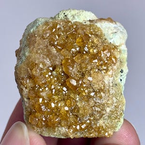 May include: A close-up of a rough, natural mineral specimen. The specimen features clusters of small, sparkling, amber-colored crystals. The base of the mineral is a light green color. The crystals are clustered together, creating a textured surface.