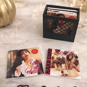 Op de afbeelding: Twee vierkante onderzetters met albumhoezen, één met de tekst "Amor Prohibido" en de andere met "Hard Core". Een zwarte krat bevat extra onderzetters. De afbeelding bevat de tekst "@Jenn.Creations1".