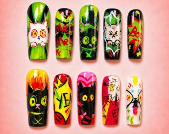 May include: A set of ten press-on nails featuring hand-painted designs with cat motifs, vibrant colors including neon green, black, red, and yellow, and text elements like Meow, Boo, and Yeah, arranged in two rows on a solid pink background.