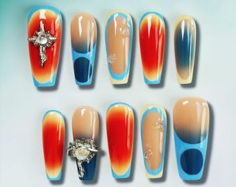 May include: A collection of ten artificial nails with diverse designs. Some nails display an orange and red gradient, while others have a nude base with blue and white details. Several nails are decorated with silver embellishments.