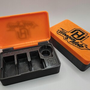 May include: An open black and orange storage box with compartments labeled 22, 20, and 10. The orange lid features the text "Hoagie Moto" and a logo. The box is designed to hold small parts or tools.
