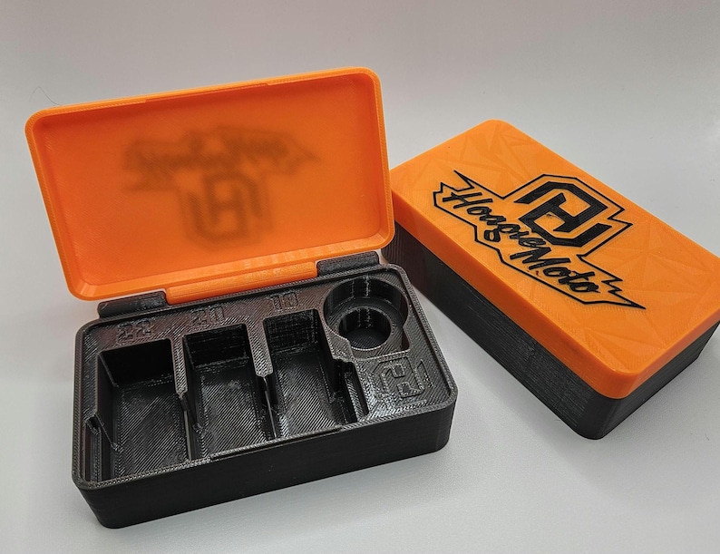 May include: An open black and orange storage box with compartments labeled 22, 20, and 10. The orange lid features the text "Hoagie Moto" and a logo. The box is designed to hold small parts or tools.