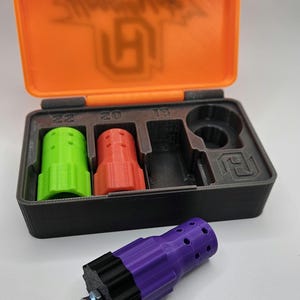 May include: An open orange and black case containing three colored cylindrical objects and a black object. A detached purple and black object with a bolt is in front of the case. The case lid has the text "Hoodlums" and a logo.
