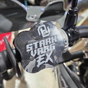 以下が含まれることがあります： 白で「STARK VARG EX」の文字が入った、黒とグレーの迷彩柄のバイクのハンドルバーエンドキャップ。キャップは黒いハンドルバーに取り付けられており、背景にはミラーやその他のバイクの部品が見えます。
