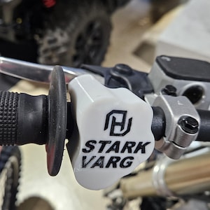 May include: A motorcycle handlebar grip with a white Stark Varg throttle control. The throttle control has the Stark Varg logo in black. The grip is black and textured.